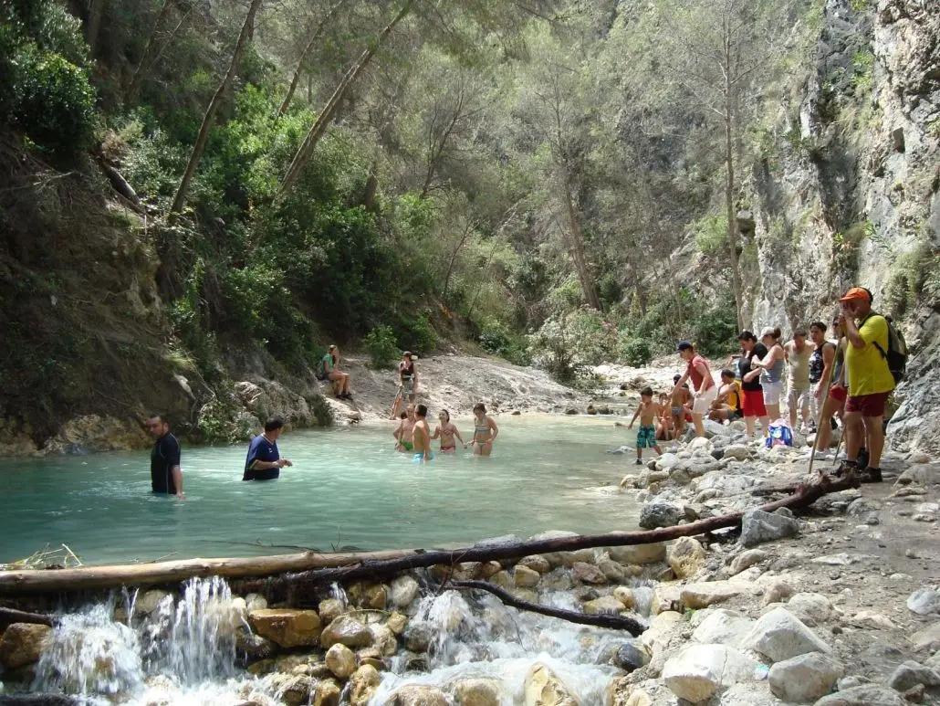 Senderistas y bañistas en una poza del río Chíllar, con un tronco cruzando el cauce