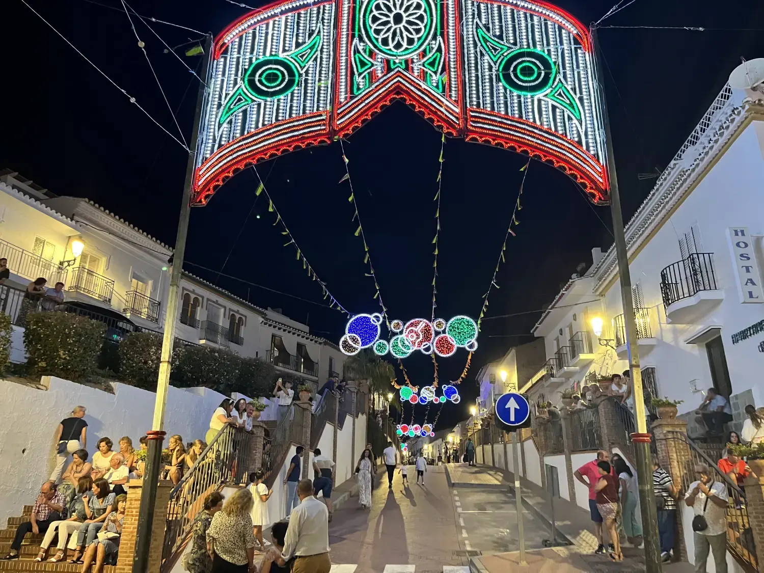 Arco de luces de colores sobre una calle en cuesta de Maro con vecinos sentados en las escaleras