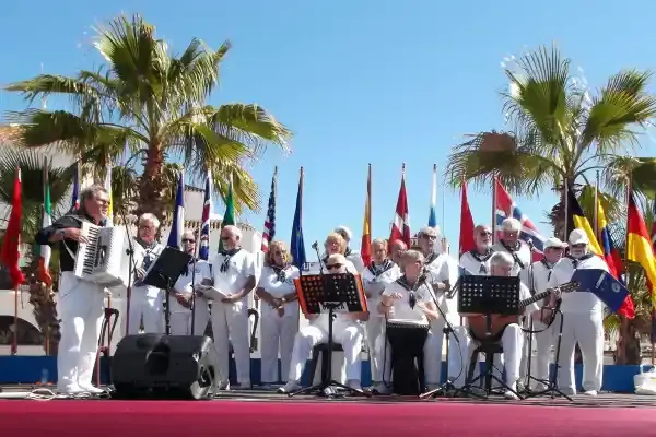 Grupo vestido de marinero cantando con acordeón y guitarra en el escenario, entre palmeras y banderas