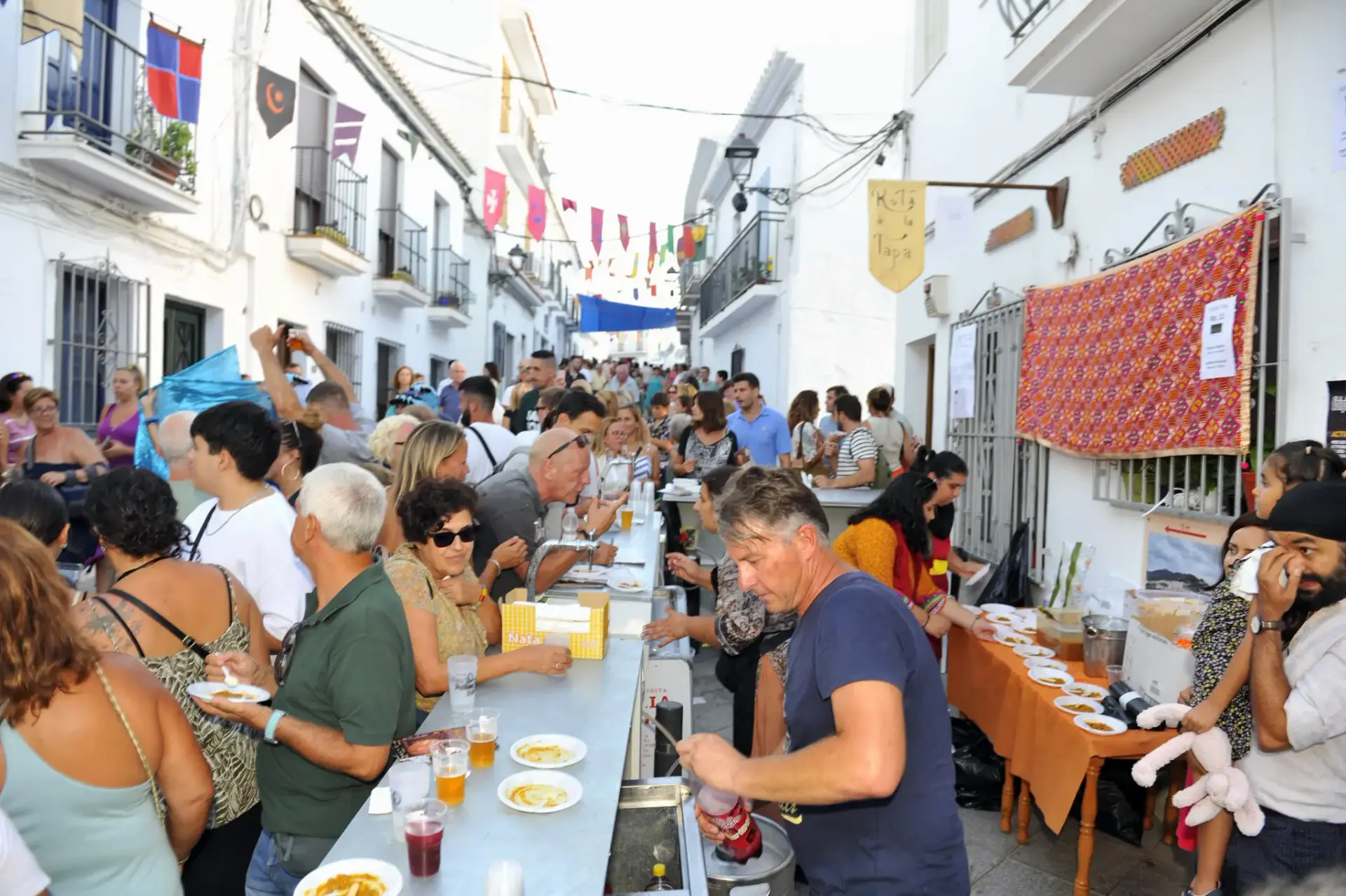 Degustación popular en mesas largas en una calle estrecha del festival, con platos servidos
