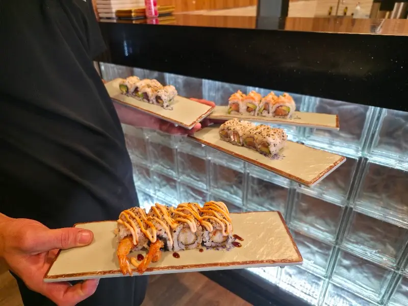 Waiter holding four trays of uramaki, one with tempura prawn and sauce