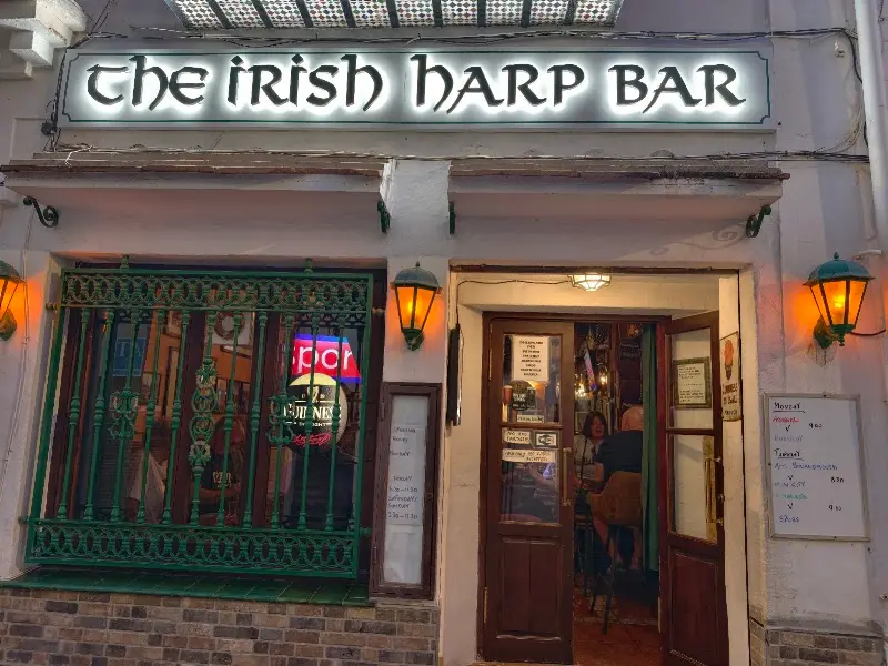 Illuminated sign of The Irish Harp Bar at night, with lanterns and the door open